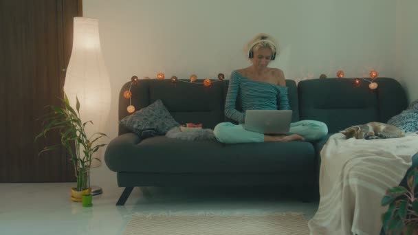 Eine Frau sitzt mit ihrem Laptop auf der Couch und hört Musik — Stockvideo