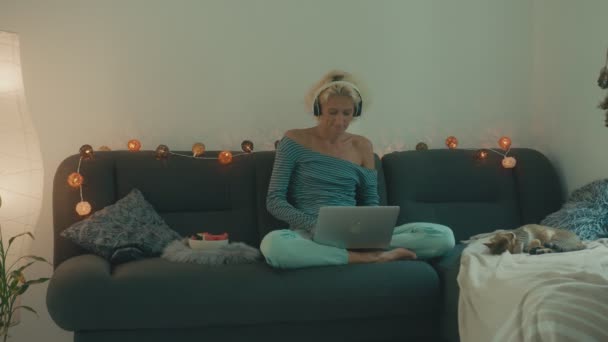 Uma mulher está sentada no sofá com seu laptop e ouvindo música — Vídeo de Stock