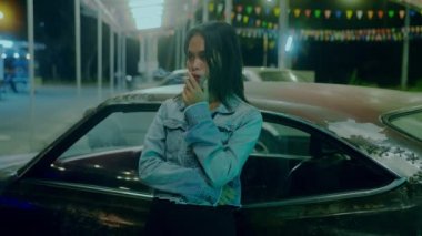 Bir kız gece geç saatlerde mesaj yazarken paslı klasik bir arabanın önünde sigara içiyor.