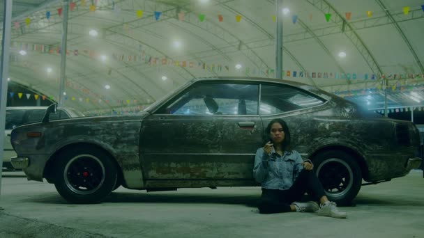 En ledsen thai flicka sitter rökning lutande mot en rostig bil på natten — Stockvideo