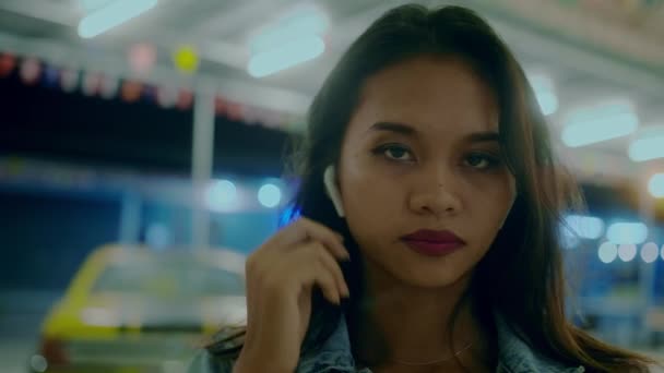A young beautiful Asian girl on the street of a night city listening to music on her smartphone headphones — Vídeo de Stock