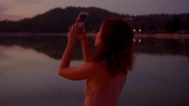 Turist kadın cep telefonuyla fotoğraf çekiyor. Paltolarında muhteşem günbatımının kamerası.