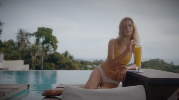 A blonde woman drinks mango shake while lounging in the sun by the pool on vacation. Elegant young woman drinks cocktail by the pool in slow motion. — Stock Video
