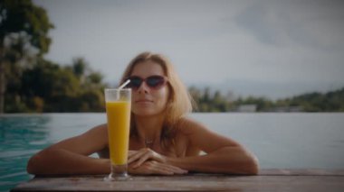 Sarışın bir kadın tatilde havuz kenarında güneşlenirken mango içkisi içer. Zarif genç bayan havuz kenarında ağır çekimde kokteyl içiyor..