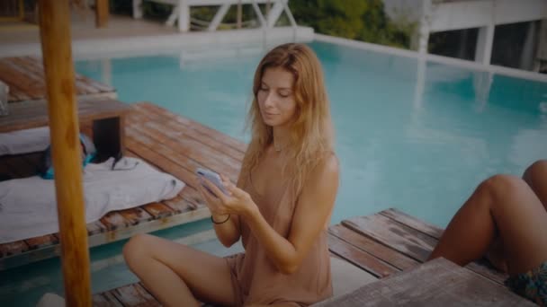 Sarışın bir kadın tatilde havuz kenarında güneşlenirken cep telefonunu kullanıyor. — Stok video
