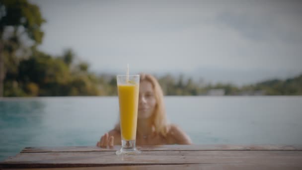 Uma mulher loira bebe um batido de manga enquanto descansa ao sol na piscina de férias. Jovem elegante bebe coquetel na piscina em câmera lenta. — Vídeo de Stock