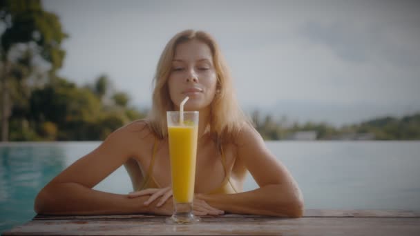 Blondynka pije koktajl mango podczas wylegiwania się na słońcu przy basenie na wakacjach. Elegancka młoda kobieta pije koktajl przy basenie w zwolnionym tempie. — Wideo stockowe