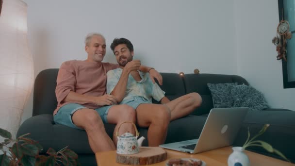 Uomini LGBTQ che guardano film su internet insieme seduti su un divano e bevono tè — Video Stock