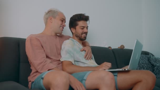 Pareja gay pasar tiempo en casa. Se acuestan en el sofá y usan su portátil. — Vídeos de Stock