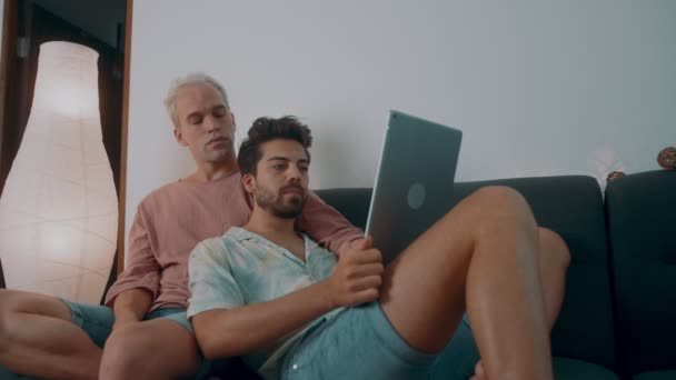 Pareja gay pasar tiempo en casa. Se acuestan en el sofá y usan su portátil. — Vídeo de stock