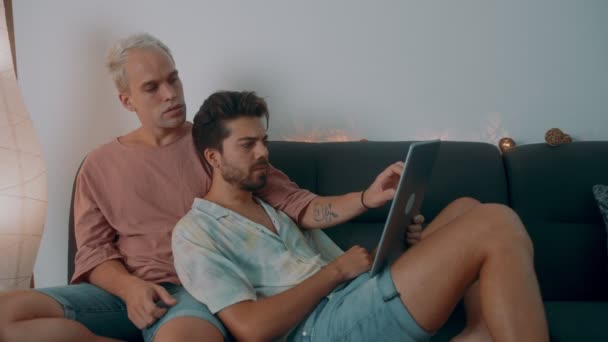 Γκέι ζευγάρι περνάει χρόνο στο σπίτι. Ξαπλώνουν στον καναπέ και χρησιμοποιούν το λάπτοπ τους.. — Αρχείο Βίντεο