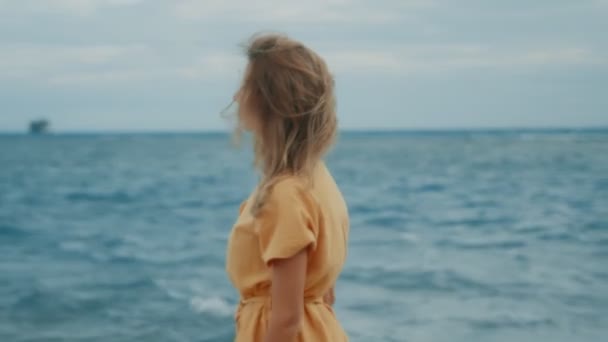 Стройная блондинка в жёлтом платье гуляет по пляжу и оглядывается вокруг — стоковое видео