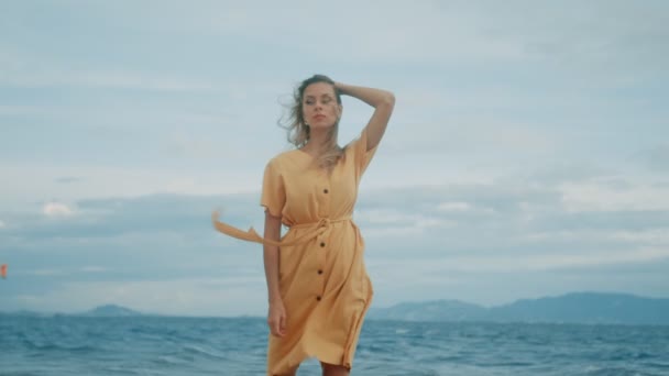 Štíhlá blondýna ve žlutých šatech kráčí podél pláže a rozhlíží se kolem — Stock video