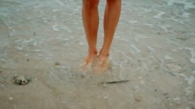 Sarı elbiseli zayıf bir sarışın kumsalda yürüyor ve etrafa bakıyor.