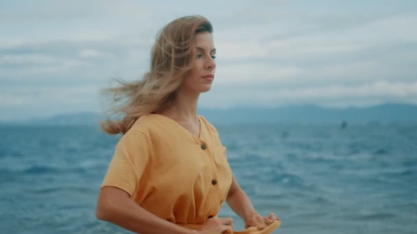 Štíhlá blondýna ve žlutých šatech kráčí podél pláže a rozhlíží se kolem — Stock video