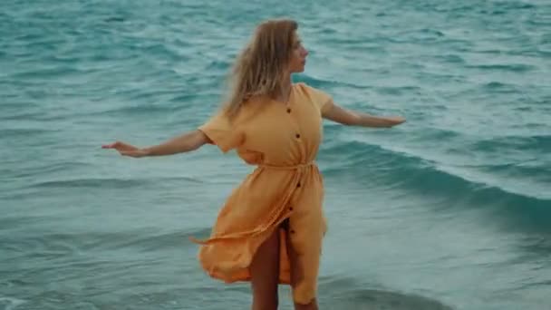 Стройная блондинка в жёлтом платье гуляет по пляжу и оглядывается вокруг — стоковое видео