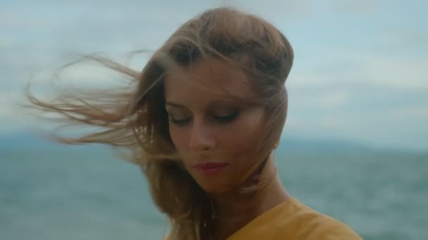 Gyönyörű szőke, sárga ruhában, egyedül a parton, ahogy a kamerát nézi felhős és szeles időben. — Stock videók