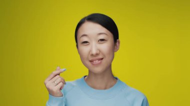 Renkli Stüdyo Çekimde Kameraya Bakan Genç Koreli Kadın Portresi. Yetişkin Japon Kız, Tek Başına Sarı Arka plan Yakından İzole Edildi. Asya Kişisi Parmaklarıyla Gülümseyen ve Kalp ya da Aşk İşareti Gösteren