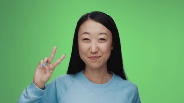 Renkli Stüdyo Çekimde Kameraya Bakan Genç Koreli Kadın Portresi. Yetişkin Japon Kız Yeşil Arkaplan 'da tek başına izole edildi. Mutlu 20 'ler Asyalı Gülümseyen ve Barış İşareti Gösteren Parmaklar