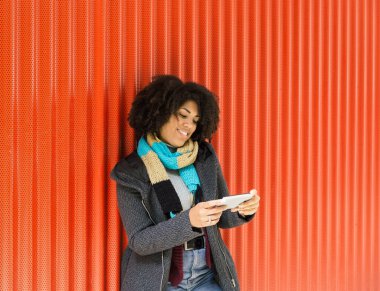 Güzel afro tarzı bir kadın, portakal rengi bir duvara yaslanırken cep telefonuna bakıp gülümsüyor.