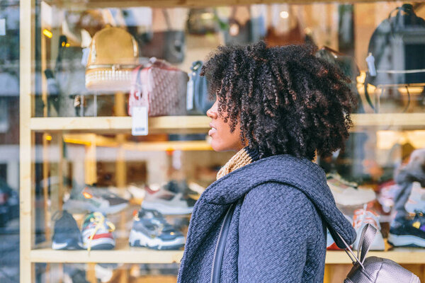 Привлекательная афроамериканская девушка в пальто и элегантно гуляет перед витриной магазина