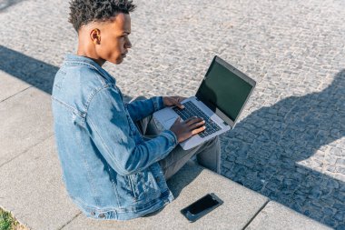 Parkta dizüstü bilgisayarı ve cep telefonunun yanında Afro saçlı adam ciddi görünüyor.