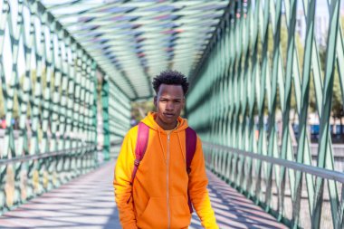 Eşofmanı ve sırt çantasıyla yeşil metal bir köprü boyunca yürüyen Afro tarzı bir çocuk.