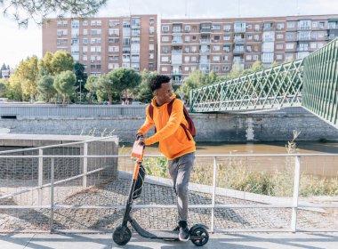 Sıradan giyinen mutlu genç adam elektrikli scooterıyla şehirde bir köprünün yanında hazırlanıyor.