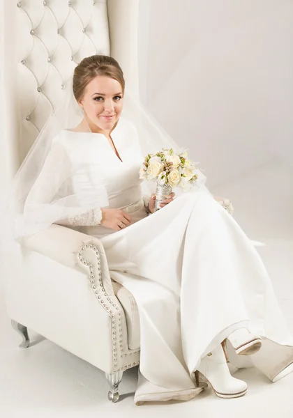 Элегантная невеста в длинном платье позирует на белом кожаном кресле — стоковое фото