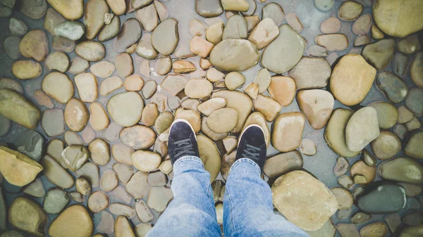Feet in sneakers standing on riverbank covered with pebbles