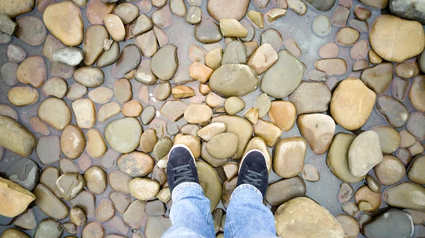 Getönter Schuss männlicher Füße in Turnschuhen, die auf Kieselsteinen im Meer stehen — Stockfoto