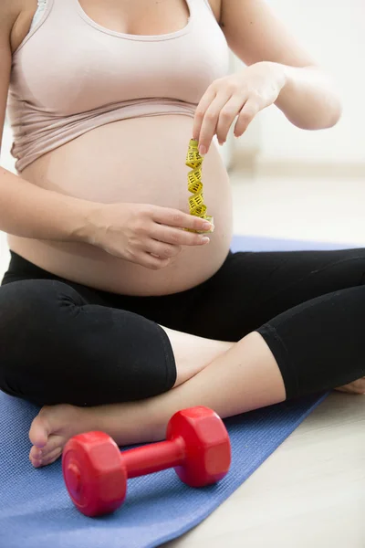 Gravid kvinna poserar med mätning av tejp och hantlar på gym — Stockfoto