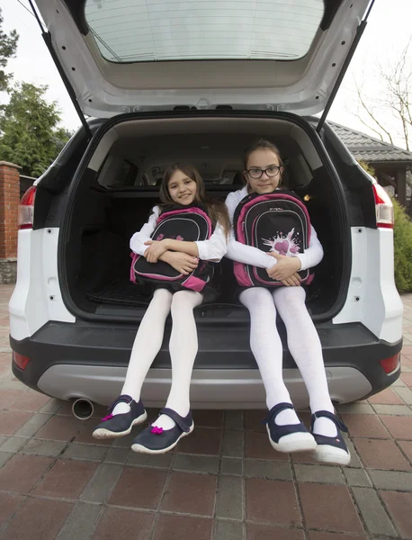 Schattig schoolmeisjes zitten in open auto kofferbak met schooltassen — Stockfoto