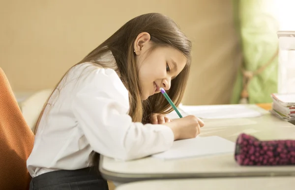 Ragazza sorridente in camicia bianca seduta dietro la scrivania e facendo homewo — Foto Stock