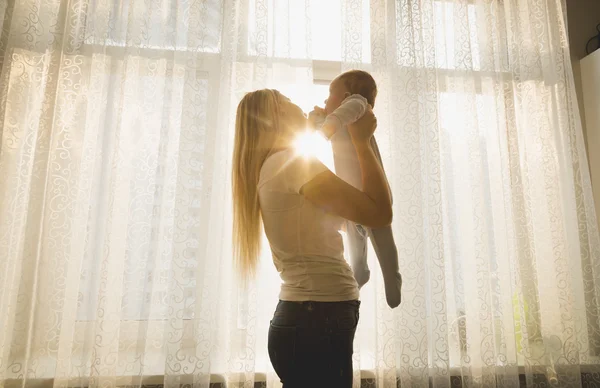 Glad mamma leker med hennes baby son mot fönstret i solen — Stockfoto
