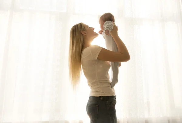 Portret van gelukkige jonge moeder opheffen van haar baby tegen windo — Stockfoto