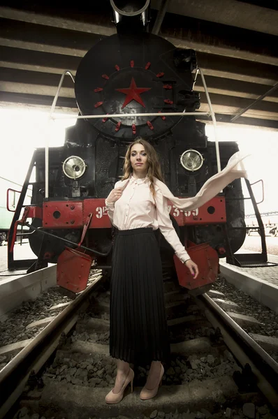 Elegant young woman standing on rails in front of old steam loco — Stock Photo, Image