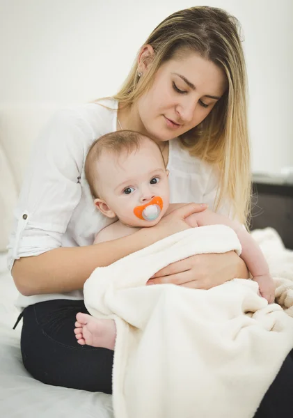 Portrait of young caring mother sitting on bed and holding baby — Stock Photo, Image