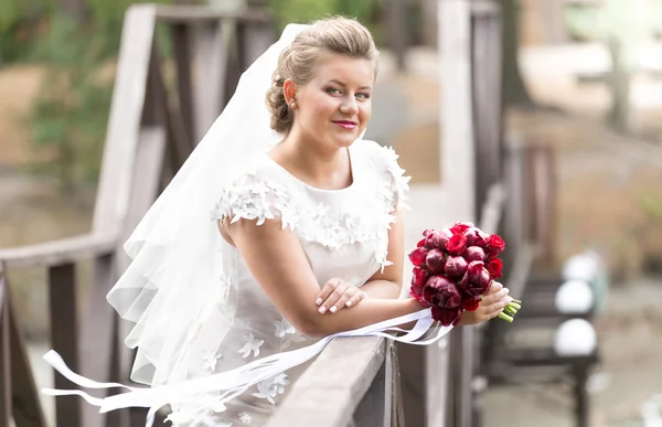 Mooie bruid met rood boeket poseren op brug op winderige dag — Stockfoto