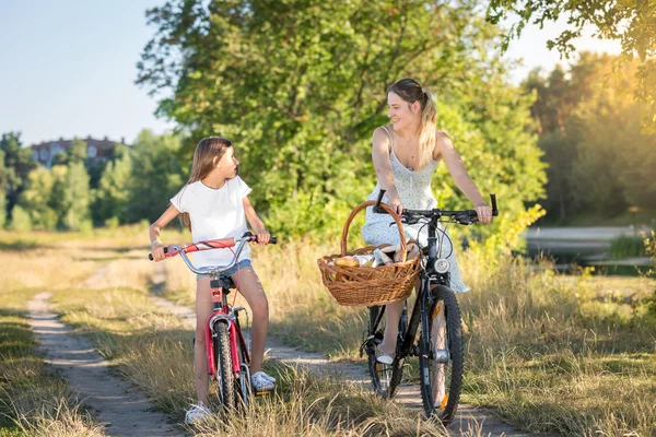 Счастливая девушка катается на велосипеде со своей матерью на лугу на закате — стоковое фото