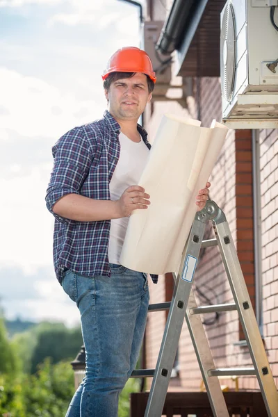 Electrician standing on high stepladder and holding plan of hous — Stock Photo, Image