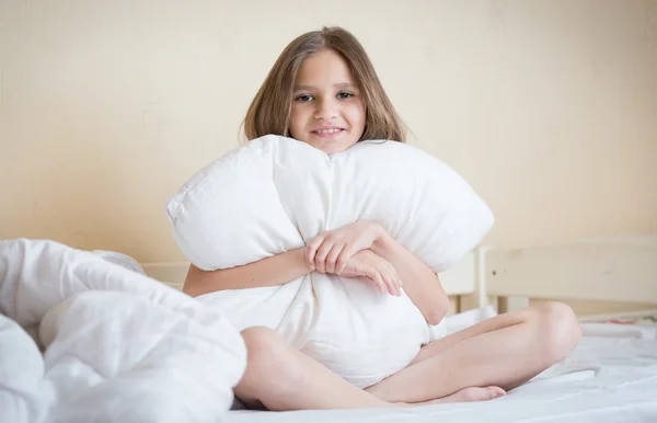 Beautiful brunette girl sitting on bed and hugging white pillow — Stock Photo, Image