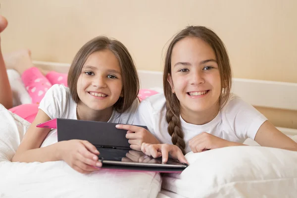 Två leende flickor ligger i sängen och använda digitala surfplatta — Stockfoto