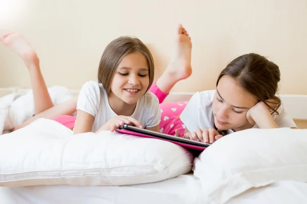 Two smiling girls lying on bed and browsing internet on digital — Stock Photo, Image