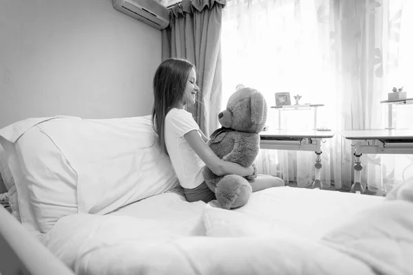 Черно-белый портрет милой девушки, сидящей на кровати и обнимающей — стоковое фото