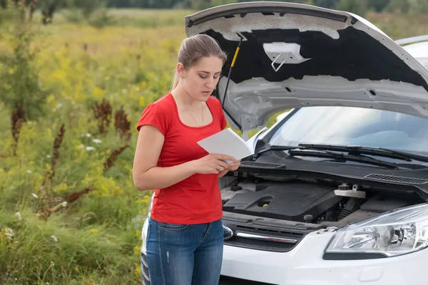 Woman standing at broken car in meadow and reading owner manual — Stock Photo, Image