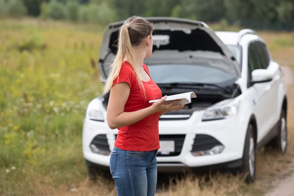 Young woman looking at her broken car and reading owner manual — Stock Photo, Image