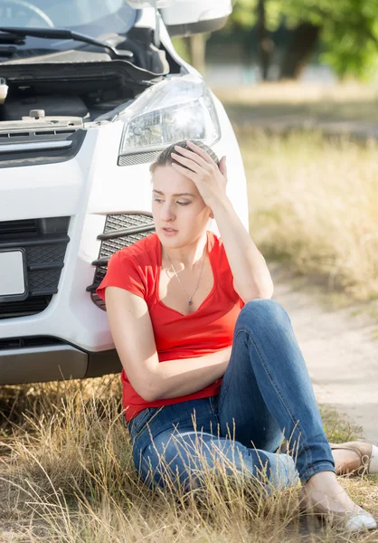 Mujer molesta sentada en el suelo y apoyada en un coche roto — Foto de Stock