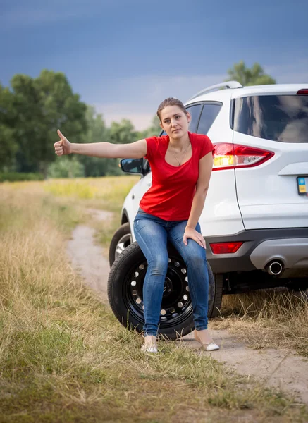 Sad woman sitting on wheel at broken car in filed and hitchhikin — Stock Photo, Image