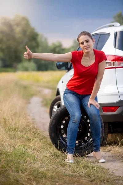 Jovem sentada em pneu sobressalente no carro quebrado e carona — Fotografia de Stock